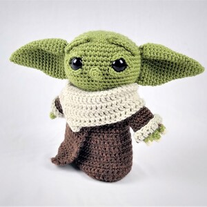 May include: A green crocheted plush toy with large eyes and a brown and white crocheted robe. The toy is a popular character from the Star Wars franchise.