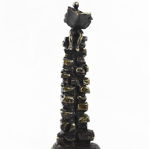 Pode incluir: Uma escultura em tom de bronze apresentando uma figura sentada no topo de uma pilha alta e estreita de blocos retangulares. A figura está posicionada no topo e parece segurar um livro aberto. A base é circular, sustentando a coluna vertical texturizada.