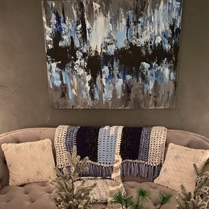 Könnte beinhalten: Abstraktes Gemälde in Blau-, Schwarz- und Grautönen, das über einem grauen Samtsofa hängt. Das Sofa ist mit zwei weißen Schneeflockenkissen und einer blau-weißen Häkeldecke ausgestattet. Kleine dekorative Bäume sind auf dem Sofa platziert.