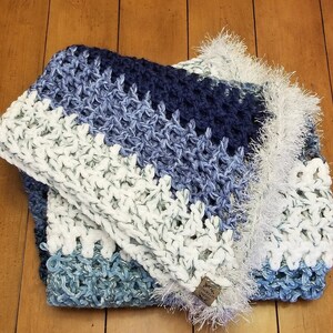 K&ouml;nnte beinhalten: Eine geh&auml;kelte Decke in Wei&szlig;, Blau und Marine. Die Decke hat einen Fransenrand und ein Etikett mit der Aufschrift "Made with Love".