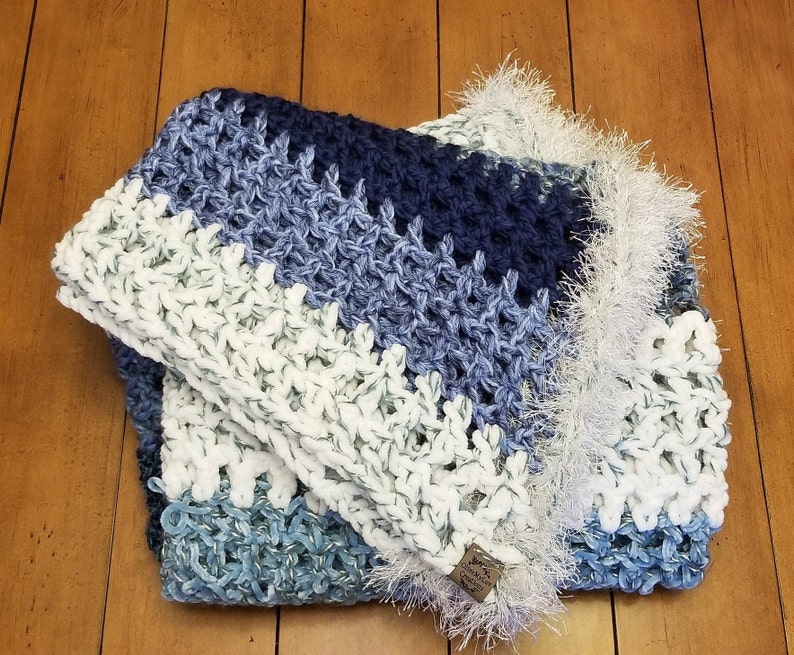 K&ouml;nnte beinhalten: Eine geh&auml;kelte Decke in Wei&szlig;, Blau und Marine. Die Decke hat einen Fransenrand und ein Etikett mit der Aufschrift "Made with Love".