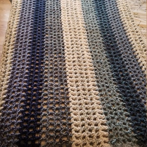 K&ouml;nnte beinhalten: Eine geh&auml;kelte Wolldecke mit einem wei&szlig;en Mittelstreifen und blauen Ombre-Streifen auf beiden Seiten. Die Decke hat einen Fransenrand.