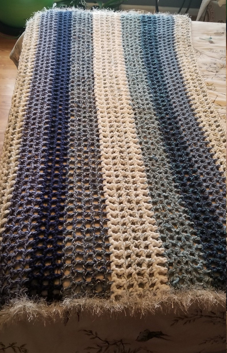 K&ouml;nnte beinhalten: Eine geh&auml;kelte Wolldecke mit einem wei&szlig;en Mittelstreifen und blauen Ombre-Streifen auf beiden Seiten. Die Decke hat einen Fransenrand.