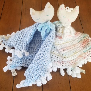 Könnte beinhalten: Zwei gehäkelte Babydecken mit angenähten Katzenspielzeugen. Eine Decke ist blau mit weißem Rand und die andere ist weiß mit rosa und grünen Streifen.