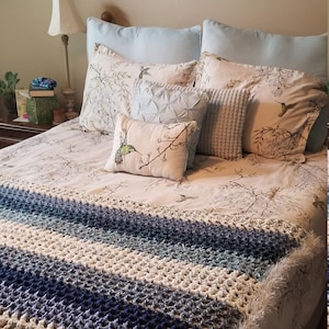 Puede incluir: Una cama blanca con una manta de crochet a rayas azules y blancas. La cama tiene una funda nórdica con estampado floral blanco y marrón y varias almohadas, incluida una almohada blanca con un diseño de colibrí.