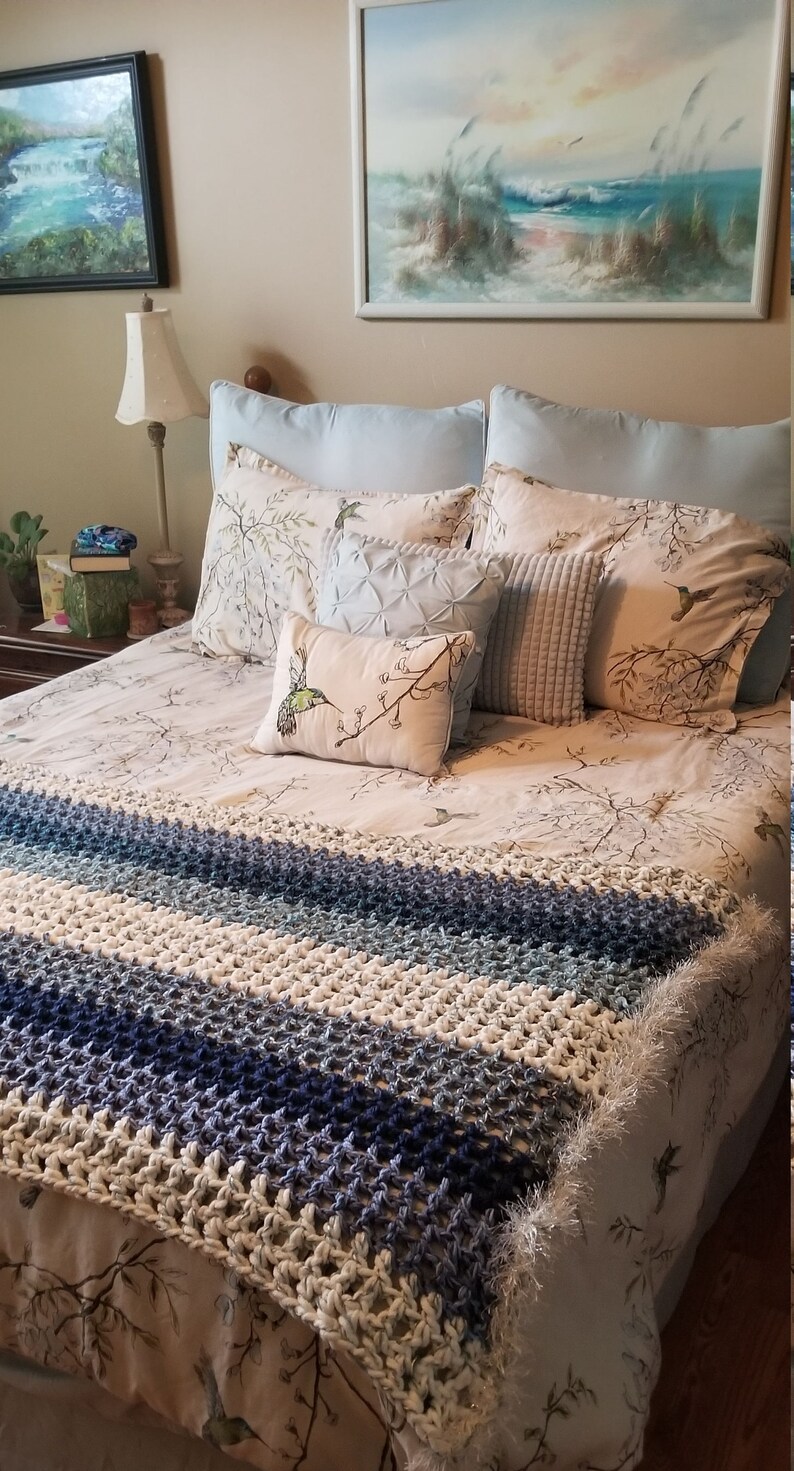 K&ouml;nnte beinhalten: Ein wei&szlig;es Bett mit einer gestreiften, geh&auml;kelten Tagesdecke in Blau und Wei&szlig;. Das Bett hat einen wei&szlig;en und braunen floralen Bettbezug und mehrere Kissen, darunter ein wei&szlig;es Kissen mit einem Kolibri-Motiv.