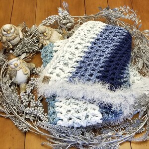 K&ouml;nnte beinhalten: Ein blauer und wei&szlig;er H&auml;kel-Schal mit einem flauschigen wei&szlig;en Saum, pr&auml;sentiert auf einem silbernen Zweigkranz mit dekorativen V&ouml;geln.