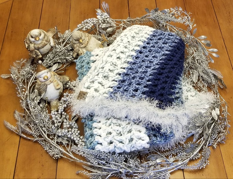K&ouml;nnte beinhalten: Ein blauer und wei&szlig;er H&auml;kel-Schal mit einem flauschigen wei&szlig;en Saum, pr&auml;sentiert auf einem silbernen Zweigkranz mit dekorativen V&ouml;geln.
