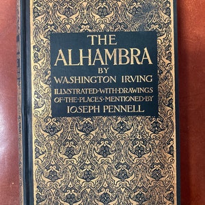 May include: A black book with gold lettering on the cover. The title is "The Alhambra" by Washington Irving. The book is illustrated with drawings of the places mentioned by Joseph Pennell.