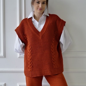 K&ouml;nnte beinhalten: Eine rostfarbene V-Ausschnitt-Strickweste mit Zopfmuster. Die Weste wird &uuml;ber einem wei&szlig;en Hemd mit Kragen getragen. Das Outfit wird durch eine passende rostfarbene Hose erg&auml;nzt. Der Gesamtstil ist l&auml;ssig und bequem.