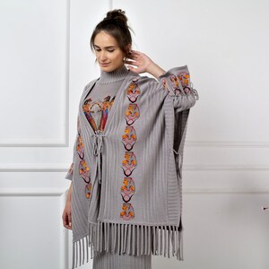 Op de afbeelding: Grijze gebreide poncho met franjes en geborduurde details. De borduursels tonen een herhalend patroon van gestileerde gezichten in oranje, rood en geel.