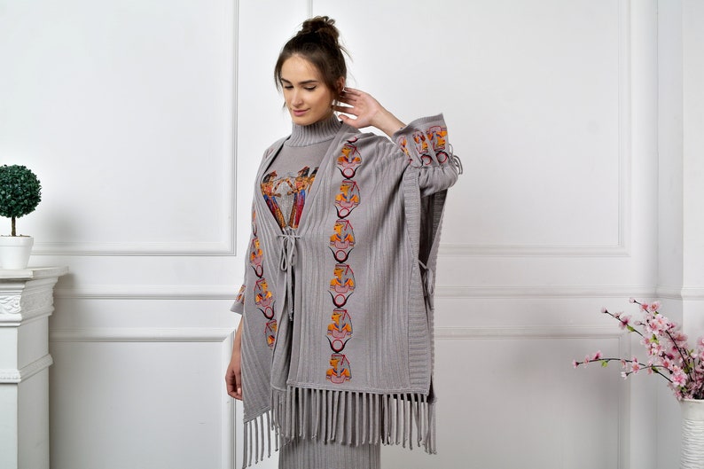 Op de afbeelding: Grijze gebreide poncho met franjes en geborduurde details. De borduursels tonen een herhalend patroon van gestileerde gezichten in oranje, rood en geel.