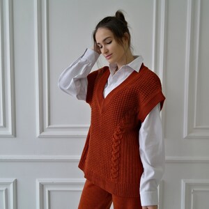 K&ouml;nnte beinhalten: Eine rostfarbene Strickweste mit V-Ausschnitt, getragen &uuml;ber einem wei&szlig;en Hemd mit Kragen. Die Weste hat ein strukturiertes Muster und ein geflochtenes Detail an der Seite. Das Outfit wird durch eine passende rostfarbene Hose erg&auml;nzt.