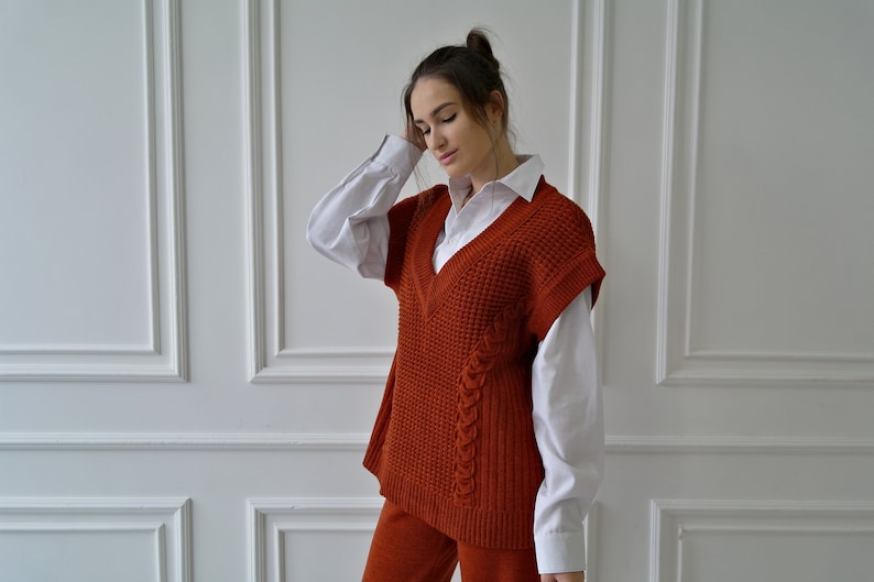 K&ouml;nnte beinhalten: Eine rostfarbene Strickweste mit V-Ausschnitt, getragen &uuml;ber einem wei&szlig;en Hemd mit Kragen. Die Weste hat ein strukturiertes Muster und ein geflochtenes Detail an der Seite. Das Outfit wird durch eine passende rostfarbene Hose erg&auml;nzt.