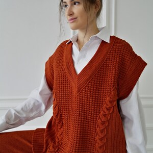 K&ouml;nnte beinhalten: Eine rostorangefarbene Strickweste mit V-Ausschnitt, Waffelstrickmuster und Zopfstrickdetails. Die Weste wird &uuml;ber einem wei&szlig;en Hemd mit Kragen getragen und mit einer passenden rostfarbenen Hose kombiniert. Das Outfit ist ein stilvolles Ensemble.