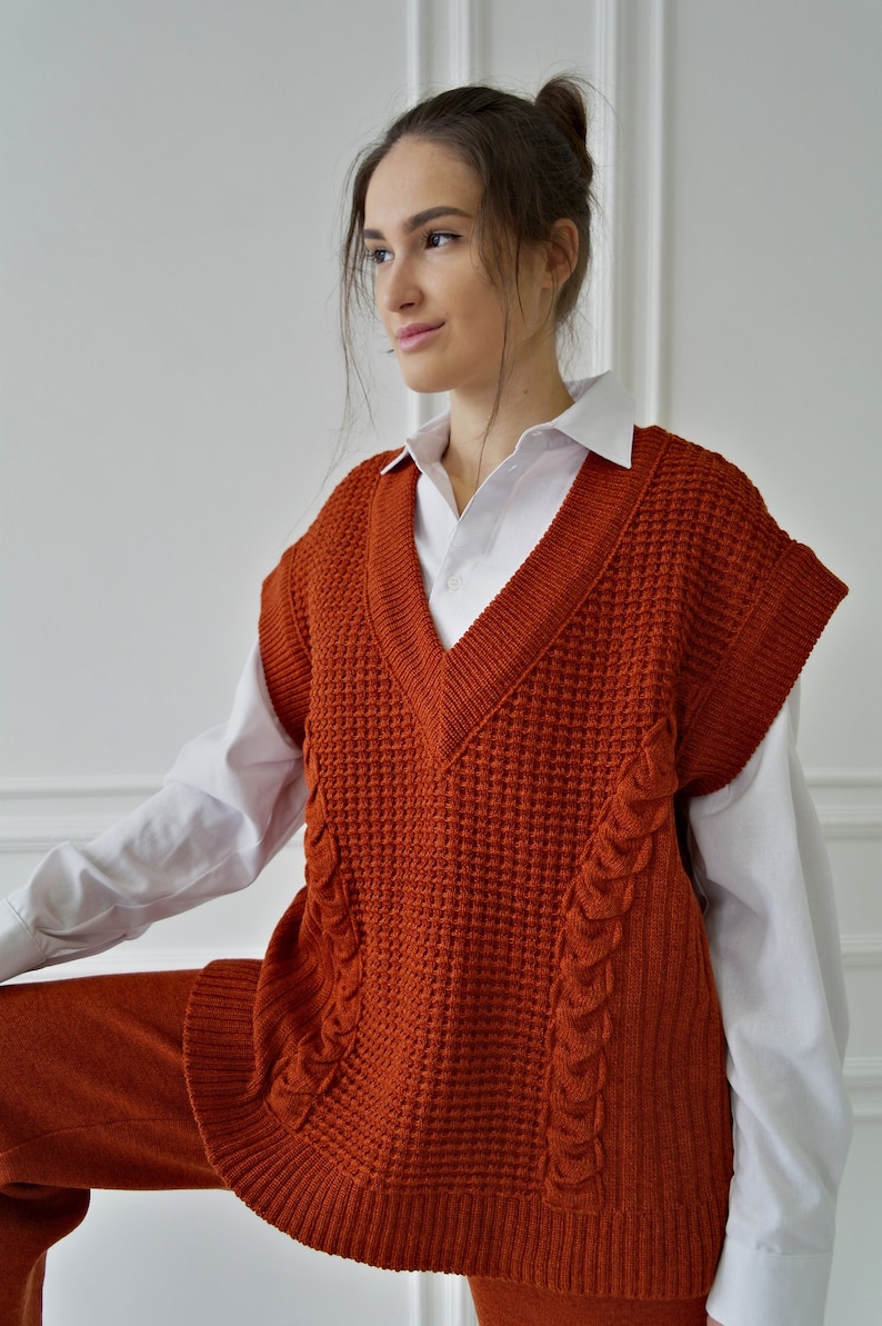 K&ouml;nnte beinhalten: Eine rostorangefarbene Strickweste mit V-Ausschnitt, Waffelstrickmuster und Zopfstrickdetails. Die Weste wird &uuml;ber einem wei&szlig;en Hemd mit Kragen getragen und mit einer passenden rostfarbenen Hose kombiniert. Das Outfit ist ein stilvolles Ensemble.