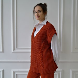 K&ouml;nnte beinhalten: Rostfarbene Strickweste mit V-Ausschnitt und Zopfmusterdetails. Die Weste wird &uuml;ber einem wei&szlig;en Hemd mit Kragen getragen. Das Outfit wird durch eine passende rostfarbene Hose erg&auml;nzt. Kleidung f&uuml;r Herbst oder Winter.