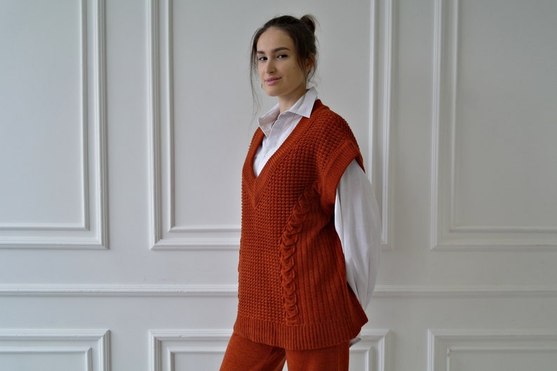 K&ouml;nnte beinhalten: Rostfarbene Strickweste mit V-Ausschnitt und Zopfmusterdetails. Die Weste wird &uuml;ber einem wei&szlig;en Hemd mit Kragen getragen. Das Outfit wird durch eine passende rostfarbene Hose erg&auml;nzt. Kleidung f&uuml;r Herbst oder Winter.