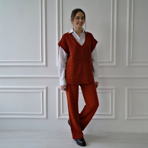 K&ouml;nnte beinhalten: Eine Frau tr&auml;gt eine rostfarbene Strickweste &uuml;ber einem wei&szlig;en Hemd mit Kragen, kombiniert mit einer passenden rostfarbenen Hose. Das Outfit wird durch schwarze Schuhe erg&auml;nzt. Der Hintergrund ist eine wei&szlig;e Wand mit dekorativen Leisten.