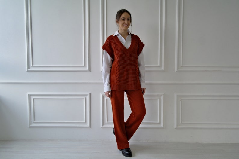 K&ouml;nnte beinhalten: Eine Frau tr&auml;gt eine rostfarbene Strickweste &uuml;ber einem wei&szlig;en Hemd mit Kragen, kombiniert mit einer passenden rostfarbenen Hose. Das Outfit wird durch schwarze Schuhe erg&auml;nzt. Der Hintergrund ist eine wei&szlig;e Wand mit dekorativen Leisten.