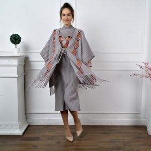 Op de afbeelding: Een grijze gebreide poncho met kleurrijke borduursels en franjes. De poncho wordt gedragen over een grijze geribbelde gebreide jurk.