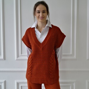 K&ouml;nnte beinhalten: Eine rostfarbene &auml;rmellose V-Ausschnitt-Strickweste mit einem strukturierten Strickmuster. Die Weste wird &uuml;ber einem wei&szlig;en Hemd mit Kragen getragen. Die Weste hat ein Zopfmuster an den Seiten. Die Person tr&auml;gt eine passende rostfarbene Hose.