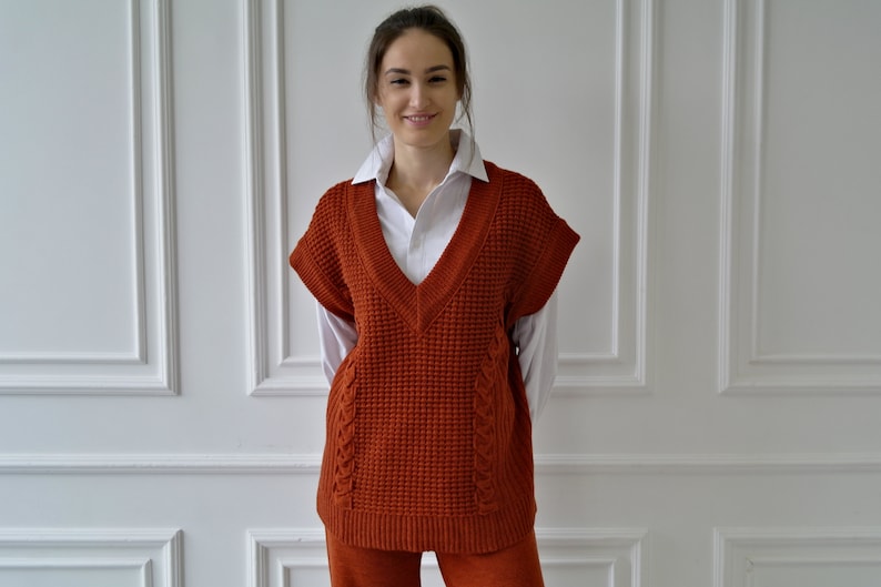K&ouml;nnte beinhalten: Eine rostfarbene &auml;rmellose V-Ausschnitt-Strickweste mit einem strukturierten Strickmuster. Die Weste wird &uuml;ber einem wei&szlig;en Hemd mit Kragen getragen. Die Weste hat ein Zopfmuster an den Seiten. Die Person tr&auml;gt eine passende rostfarbene Hose.