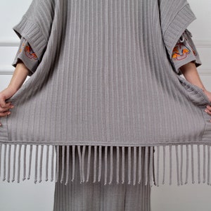 Op de afbeelding: Een grijze gebreide cardigan met een franje rand. De cardigan heeft geborduurde details op de mouwen.