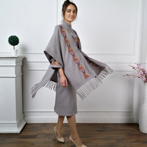 Op de afbeelding: Een grijze gebreide poncho met een geborduurd bloemenmotief en franjes. De poncho wordt gedragen over een bijpassende grijze gebreide jurk.