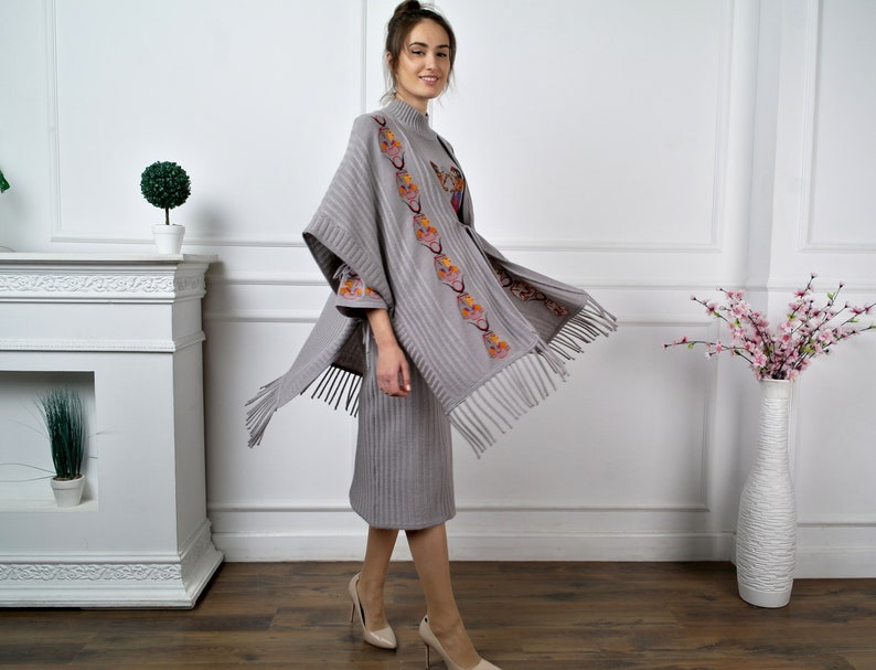 Op de afbeelding: Een grijze gebreide poncho met een geborduurd bloemenmotief en franjes. De poncho wordt gedragen over een bijpassende grijze gebreide jurk.