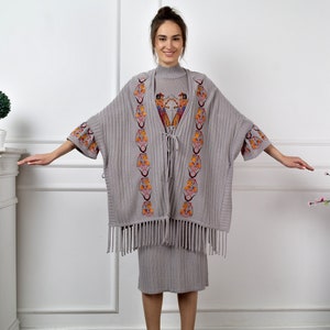 Op de afbeelding: Een grijze gebreide poncho met kleurrijke geborduurde details en franjes. De poncho wordt gedragen over een grijze geribbelde jurk.