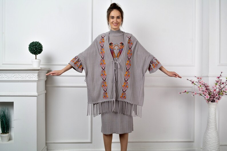Op de afbeelding: Een grijze gebreide poncho met kleurrijke geborduurde details en franjes. De poncho wordt gedragen over een grijze geribbelde jurk.