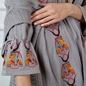 Op de afbeelding: Grijze gebreide cardigan met geborduurde ontwerpen ge&iuml;nspireerd op Egypte. De cardigan heeft een lange, losse pasvorm en wijde mouwen met strikjes aan de manchetten.