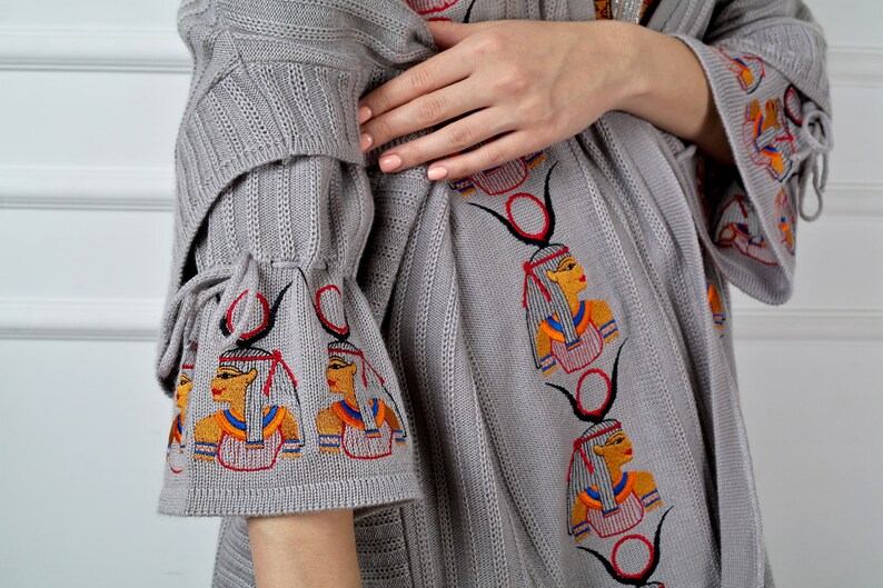 Op de afbeelding: Grijze gebreide cardigan met geborduurde ontwerpen ge&iuml;nspireerd op Egypte. De cardigan heeft een lange, losse pasvorm en wijde mouwen met strikjes aan de manchetten.