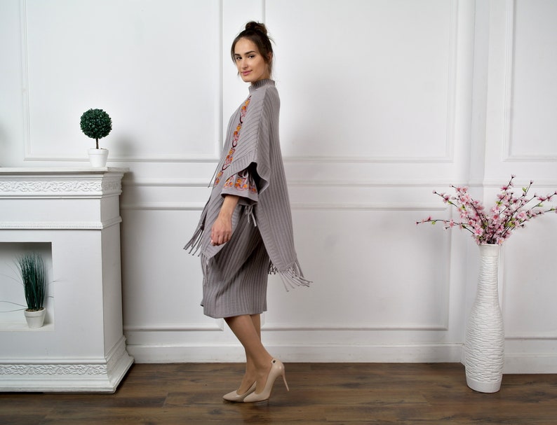 Op de afbeelding: Een grijze poncho met franjes en geborduurde details. De poncho wordt gedragen door een persoon die in een kamer met witte muren en een open haard staat.