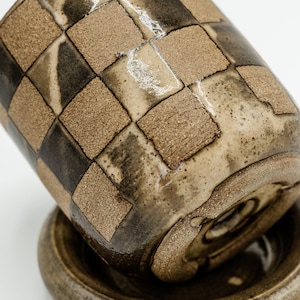 May include: A handmade ceramic cup and saucer set. The cup features a checkerboard pattern in shades of brown and black. The saucer is a matching brown color. The set is designed with a rustic, artisanal aesthetic.