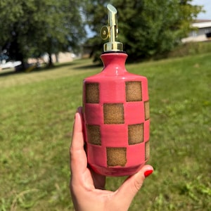 Peut inclure: Un distributeur d'huile en céramique avec une base rose vif ornée d'un motif répétitif de carrés bruns texturés. La bouteille est surmontée d'un bec verseur de couleur or métallique, offrant une fermeture fonctionnelle pour le récipient.