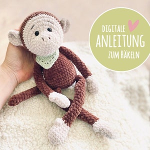 Op de afbeelding: Een bruin gehaakt aapje met een groen gehaakt slabbetje. De aap zit op een wit, pluche oppervlak. De tekst "DIGITALE ANLEITUNG ZUM HÄKELN" staat in een cirkel met een roze hart.