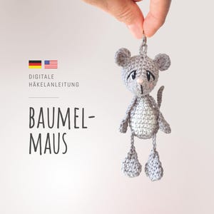 Könnte beinhalten: Ein gehäkeltes graues Mäuse-Schlüsselanhänger mit weißem Bauch, rosa Nase und schwarzen Augen. Die Maus wird von einem silbernen Schlüsselring gehalten. Das Bild enthält den Text "BAUMEL-MAUS" und "DIGITALE HÄKELANLEITUNG".