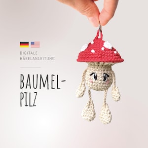 Puede incluir: Un llavero de hongo tejido a ganchillo con un gorro rojo y lunares blancos, tallo beige y piernas colgantes. El hongo tiene una cara sonriente con ojos negros y mejillas rosadas. La imagen incluye el texto "BAUMEL-PILZ" y "DIGITALE HÄKELANLEITUNG".