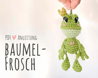 May include: A green crocheted frog wearing a yellow crown with pink cheeks and a pink heart on its chest. The frog is attached to a silver key chain. The text "PDF ♥ ANLEITUNG BAUMEL-FROSCH" is in the image.