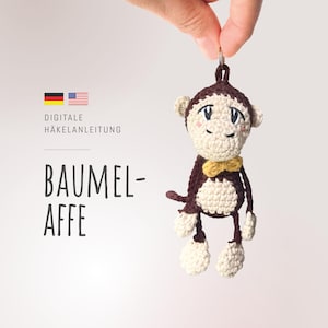 Puede incluir: Un llavero de mono tejido a crochet con pelaje marrón, una pajarita amarilla y pies blancos. El mono está colgado de un anillo plateado. El texto "Digitale Häkelanleitung" y "Baumel-Affe" es visible en la imagen.
