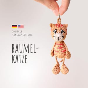May include: A small, light brown crocheted cat figurine with darker brown stripes, a white muzzle, and embroidered facial features. The cat has a loop at the top for hanging. Text on the image reads: DIGITALE HÄKELANLEITUNG BAUMEL-KATZE.