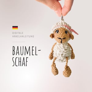 Könnte beinhalten: Ein gehäkeltes weißes Schaf mit braunen Augen und einer rosa Nase, das an einem silbernen Schlüsselring hängt. Das Schaf ist ein Schlüsselanhänger und ist mit "Digitale Häkelanleitung Baumel-Schaf" beschriftet.