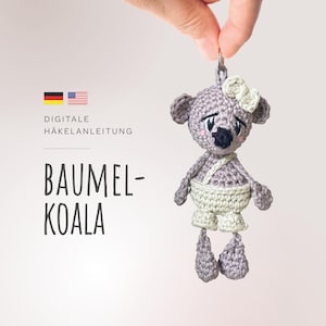 Könnte beinhalten: Ein gehäkelter Koala-Schlüsselanhänger mit einer rosa Schleife, der an einer silbernen Kette hängt. Der Koala trägt eine weiße gehäkelte Windel. Der Text "Digitale Häkelanleitung" und "Baumel-Koala" ist im Hintergrund sichtbar.