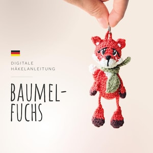 May include: A crocheted fox keychain with a green scarf. The fox is red with white accents and brown feet. The keychain is labeled "Digitale Häkelanleitung Baumel-Fuchs".