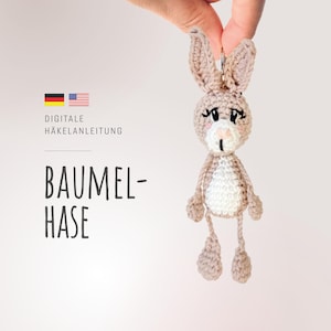 May include: A crocheted bunny rabbit keychain in beige and white with black eyes and a pink nose. The bunny is hanging from a silver key ring. The text "Digitale Häkelanleitung" and "Baumel-Hase" is visible in the background.
