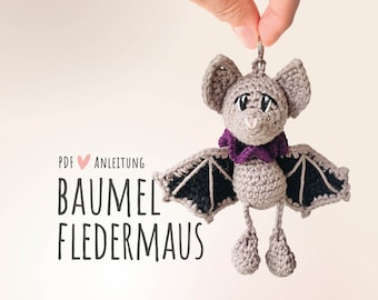 Puede incluir: Un juguete de peluche de murciélago de ganchillo con un collar morado, alas negras y un corazón rosa. El texto "PDF ♥ ANLEITUNG BAUMEL FLEDERMAUS" es visible en la imagen.