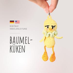 Könnte beinhalten: Ein gelber gehäkelter Küken-Schlüsselanhänger mit grünem Rüschenkragen und schwarzen Augen. Der Schlüsselanhänger ist mit "BAUMEL-KÜKEN" und "DIGITALE HÄKELANLEITUNG" beschriftet.