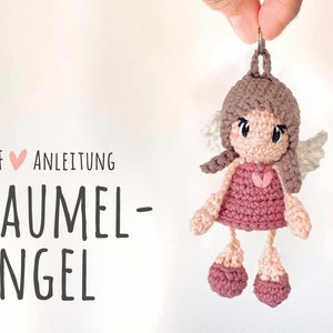 Könnte beinhalten: Ein gehäkelter Engel-Schlüsselanhänger aus rosa und braunem Garn. Der Engel trägt ein rosa Kleid, hat braunes Haar und weiße Flügel. Der Schlüsselanhänger ist an einem silbernen Ring befestigt.